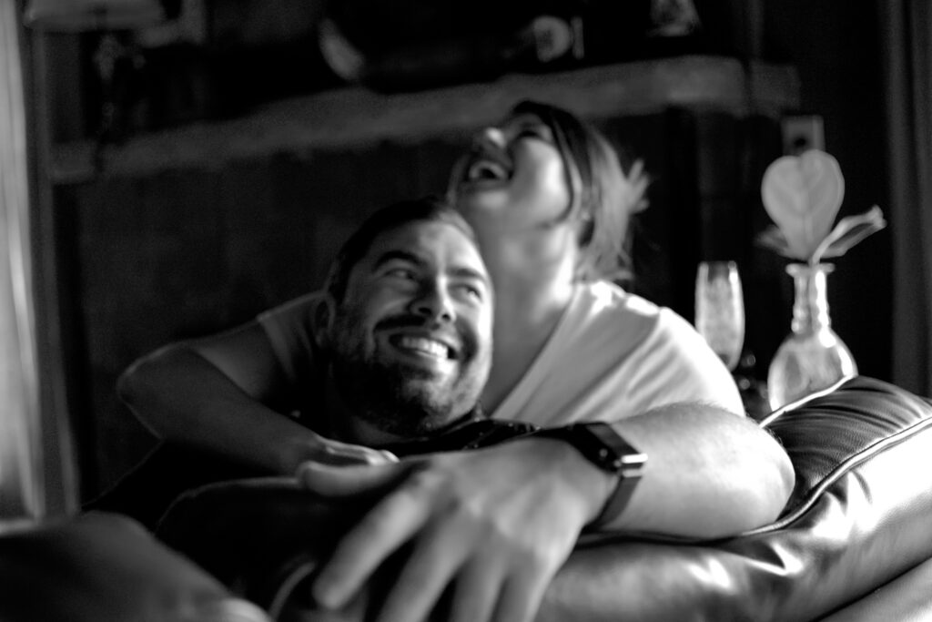 This is a photo by Kat Stano of a cute couple laughing on their couch.