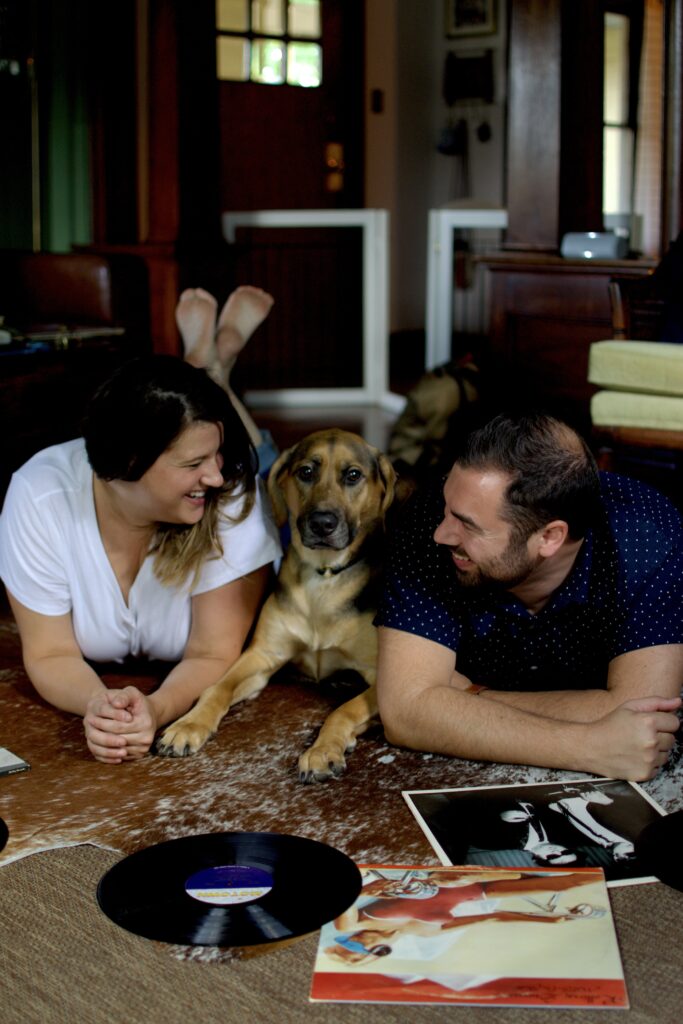 This is a photo by Kat Stano of a cute couple sitting on the floor in their home with their dog, enjoying their record player's records.