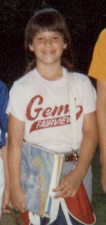 This is a childhood photo of Kat Stano in her Gems softball team uniform, holding a bag and a unicorn Trapper Keeper.