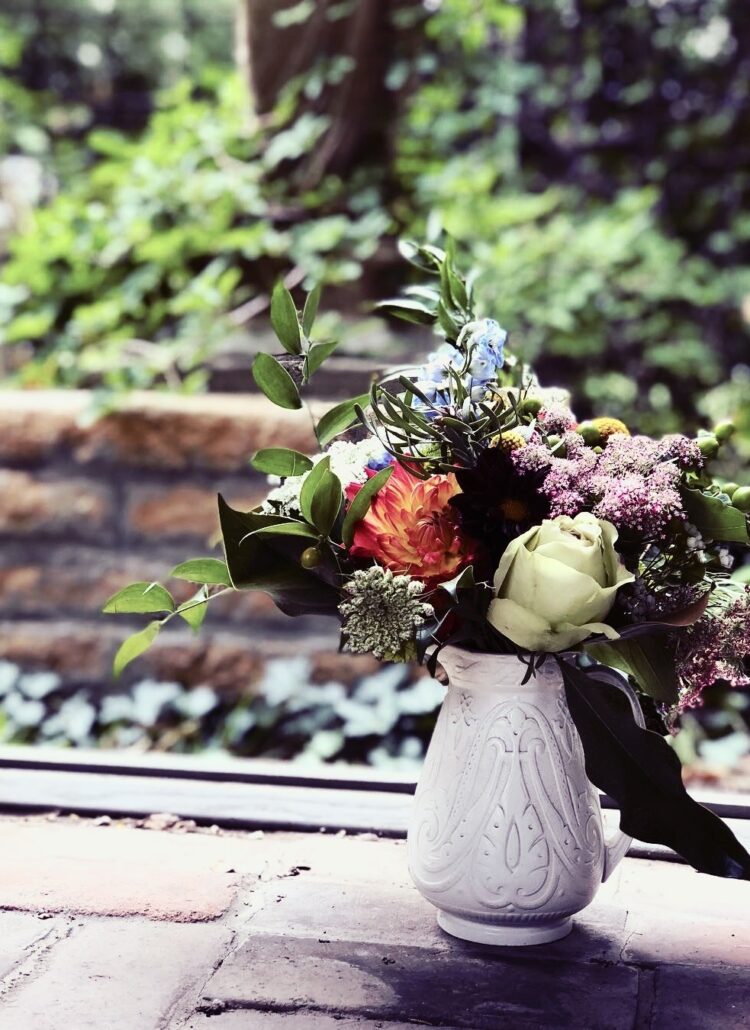 Beautiful flowers in a decorative pitcher, sitting on a brick path in a garden. Photo taken by Kat Stano.