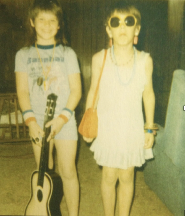 This is a childhood photo of Kat and her brother dressed up in each other's clothes with jewelry, a purse, sunglasses, workout wristbands, and a guitar.