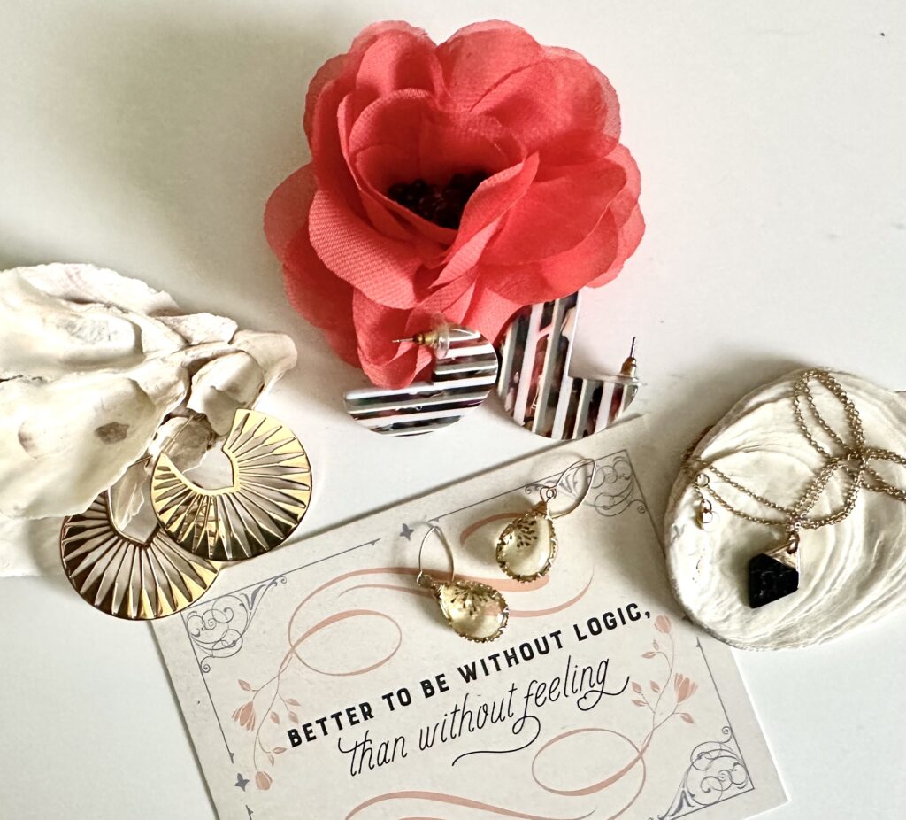 This is a photo by Kat Stano of a jewelry flat lay with three pairs of earrings, a necklace, two seashells, a flower, and a Jane Austen quote that says, "Better to be without logic than without feeling."