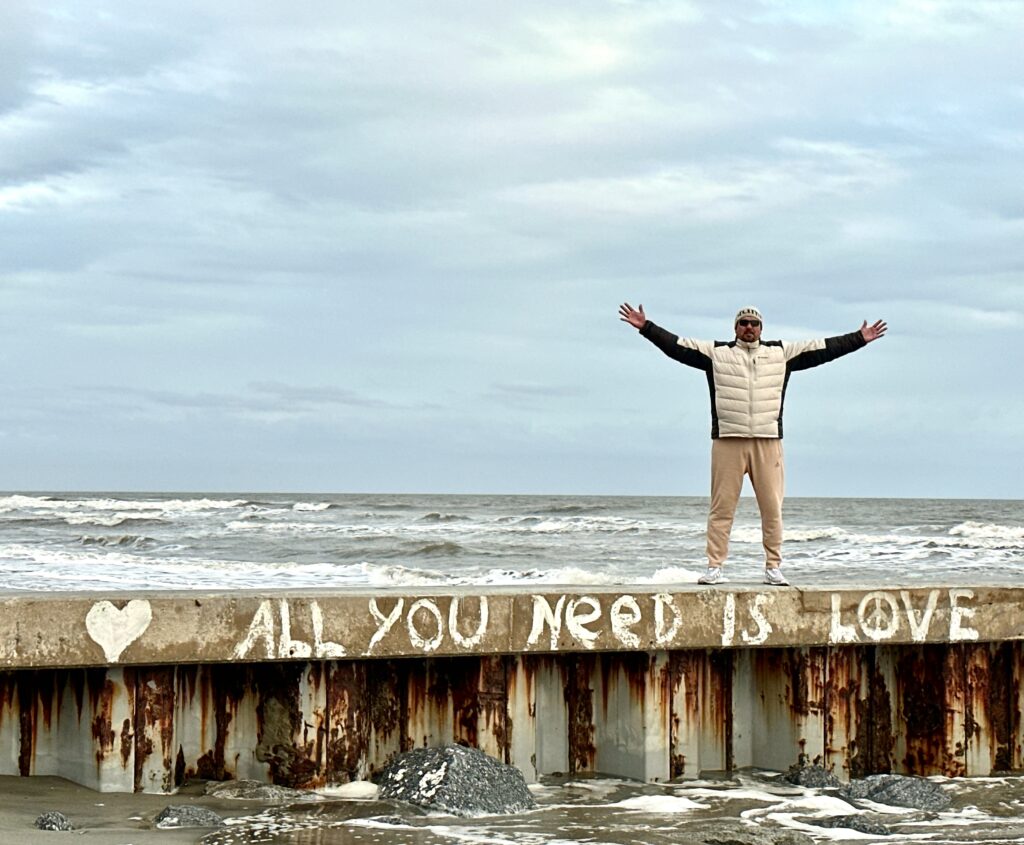 This is a photo by Kat Stano of her husband standing on a platform in front of the ocean at Folly Beach, South Carolina. The platform reads, "All you need is love."