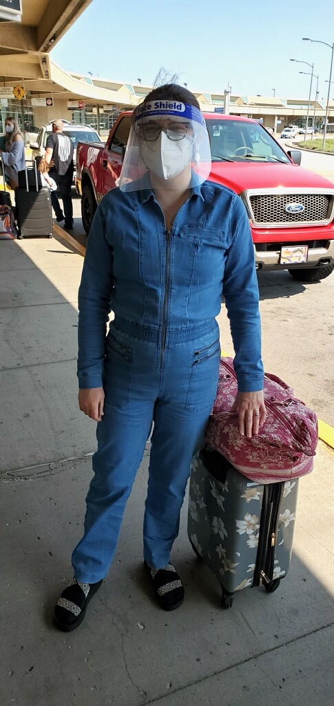 This is a photo of Kat Stano standing at the airport with a face mask, face shield, and luggage.