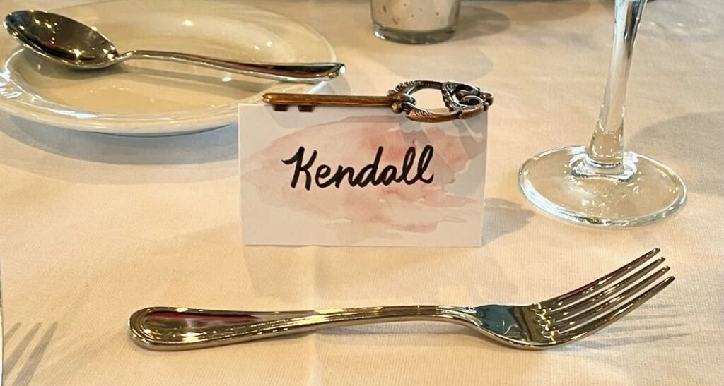 This is a photo of a wedding place card holder with a skeleton key bottle opener attached on a dinner table with silverware, a plate, and wine glass.