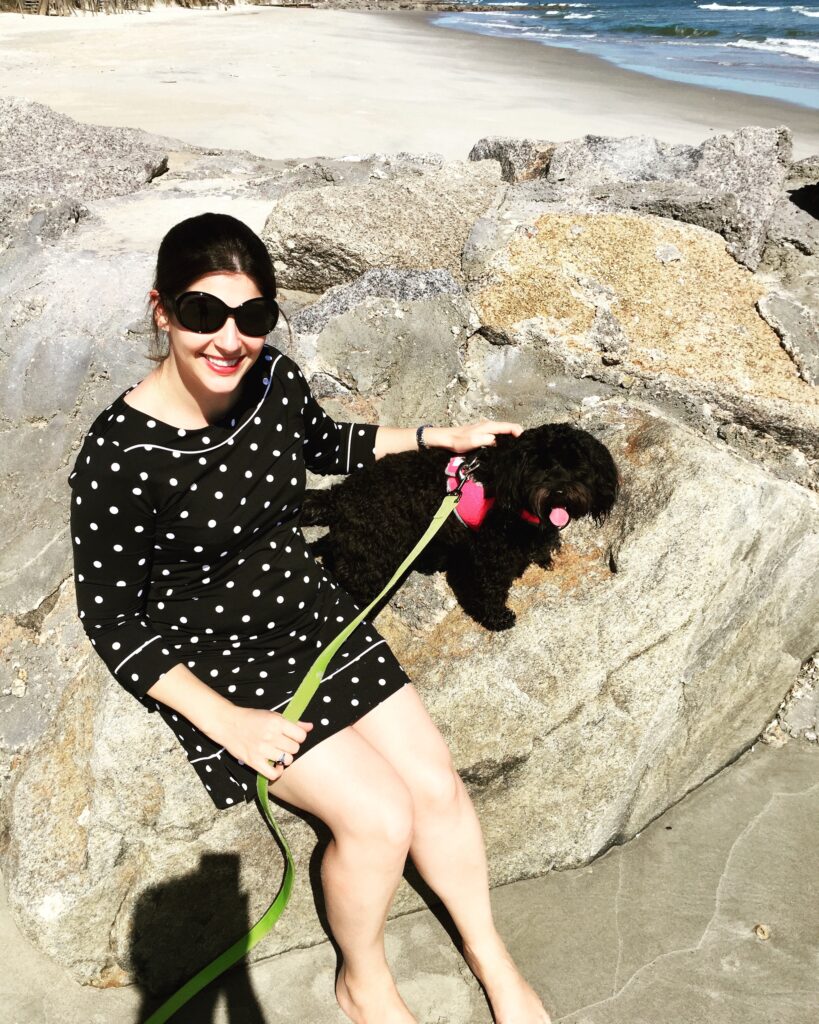 This is a photo of Kat Stano sitting on a rock at the beach with her schnoodle dog, Cosette.