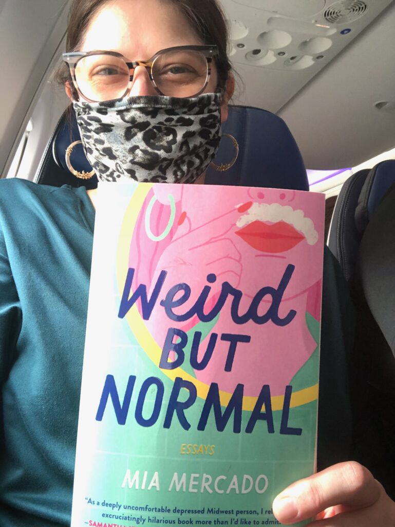 This is a photo of Kat Stano on a plane, wearing a face mask and holding a book by Mia Mercado titled, "Weird But Normal."