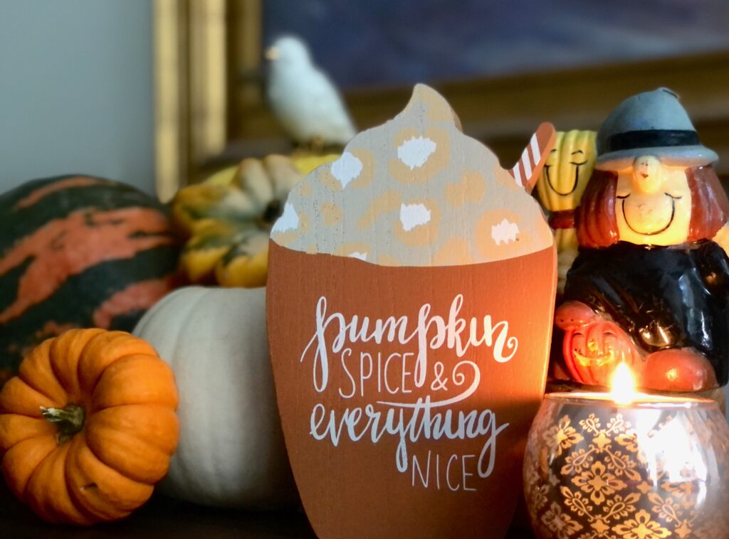 This is a photo of an autumn display with pumpkins, a friendly witch figurine, a candle, a bird, and a sign that says, "Pumpkin spice and everything nice."