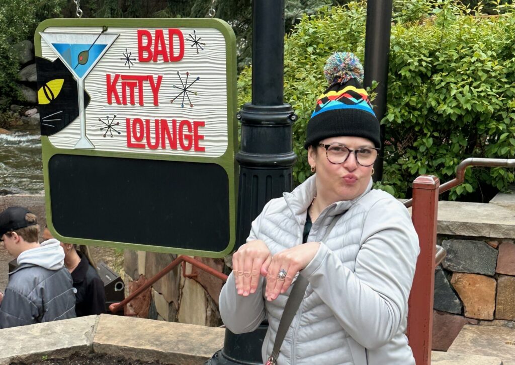 This is Kat in front of a sign that says Bad Kitty Lounge, while wearing a pom-pom beanie as a reminder of holiday travel safety tips to bring warm clothes.