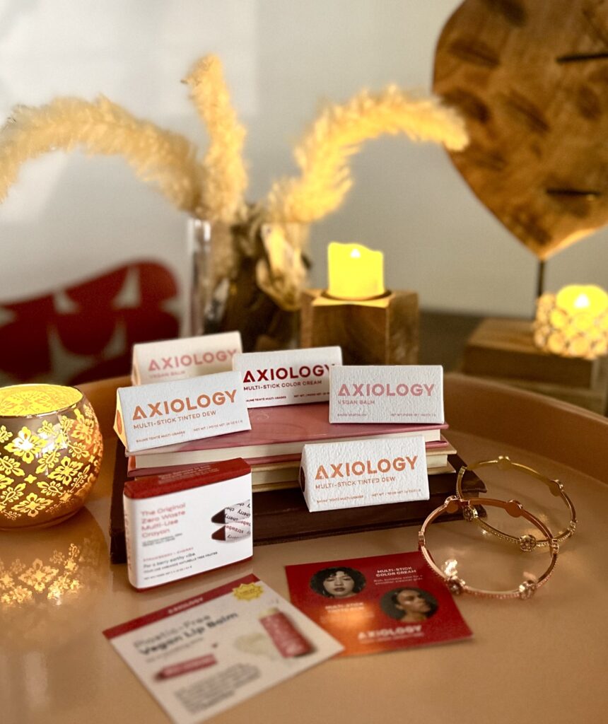 This is a photo of Axiology Balmies, multi-sticks, and tinted dews to illustrate a great zero-waste makeup brand. Also included are candles, pampas flowers, heart statue, seashells, books, and bracelets.