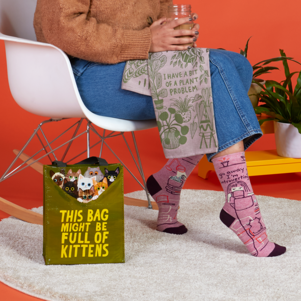 This is a photo by Blue Q with a girl sitting by a bag that says, "This bag might be full of kittens" and holding a dishtowel that says, "I have a bit of a plant problem" and wearing socks that say, "Go away I'm introverting" which represents gifts for environmental activists.