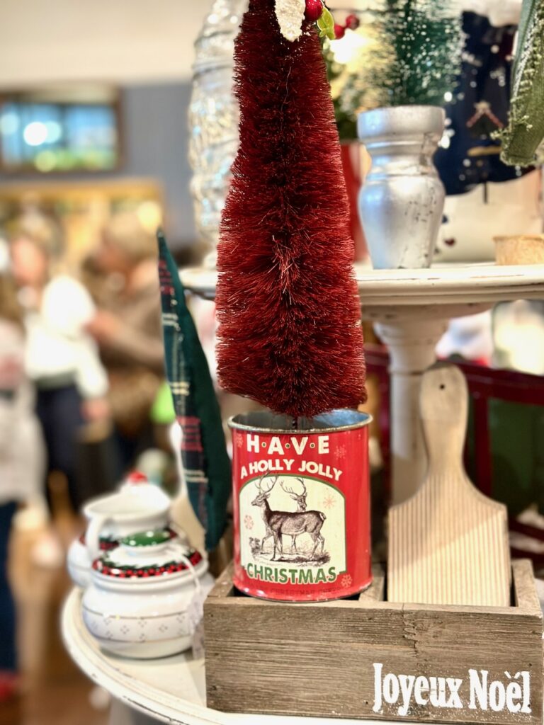 This is a canned food aluminum can decorated with a reindeer label that says, "Have a holly jolly Christmas" used as a vase for a crimson bottle brush tree to describe holiday decorating tips.