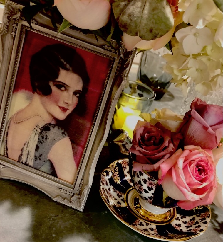 This is a vintage table display from Kat's wedding of a flapper girl print, roses in a teacup, flowers, and candles to describe sustainable wedding favors reddit.