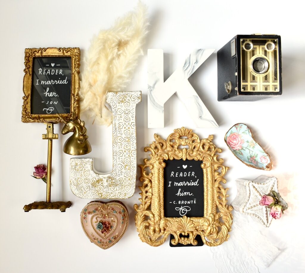 This is a flatlay of items from Kat's wedding with quotes that say, "Reader, I married him" and "Reader, I married her", a unicorn bell, dried flowers, pampas grass, J and K initials, a heart box, an antique camera, an oyster shell, and trinket box to show you have a kind heart meaning.