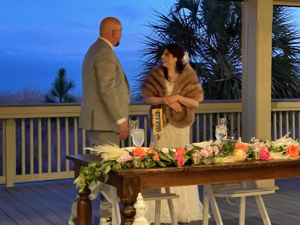 This is Jon and Kat at their wedding in front a farmhouse table overlooking the ocean, with colorful florals, candles, and quotes to show eco friendly wedding centerpieces.