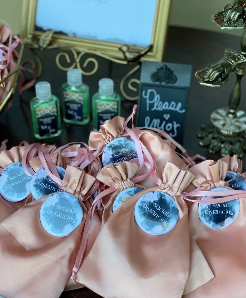 This is a photo of Kat's wedding with masks in rose gold pouches, social distancing buttons, and customized hand sanitizer with a "please enjoy" sign and antiques to discuss wedding exit.