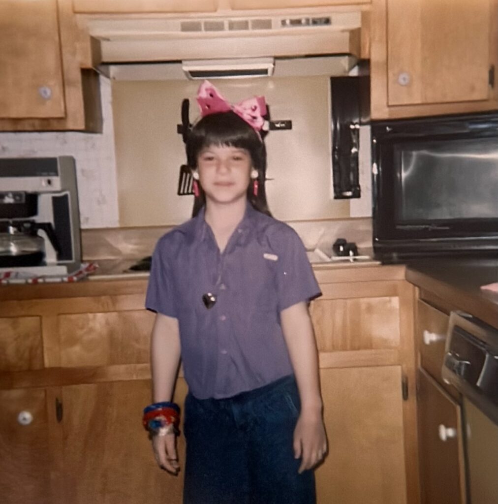 This is Kat as a child in the 1980s, standing in the kitchen by the stove and coffee pot, wearing a pink bow with rhinestones, hot pink and white clip-on earrings, a gold heart necklace, purple shirt, jeans, and a bunch of bangles to add to the story of business card holder ideas.