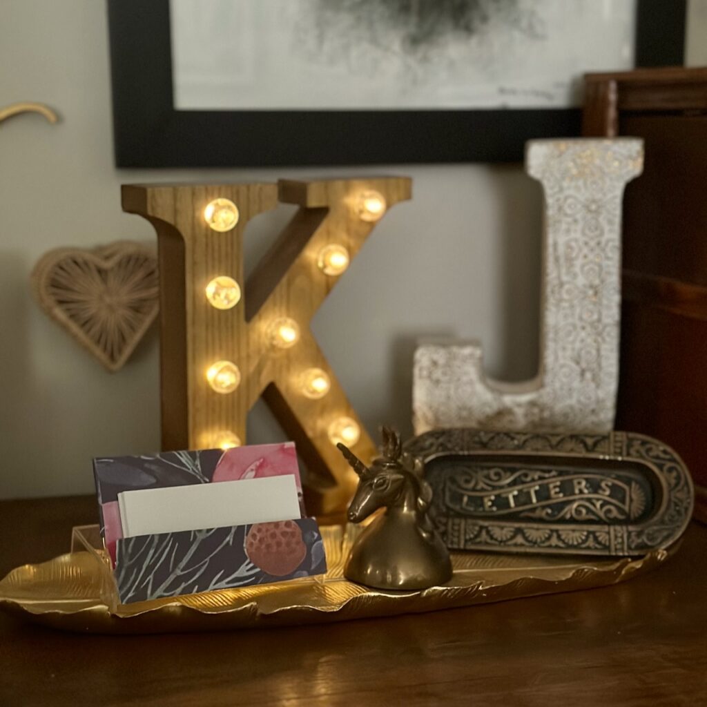 This is a table with a decorative vignette with a diy cute business card case made out of a cassette tape holder and it's holding business cards; also a black and white illustration is framed on the wall, and also on the table a wooden letter "K" with lights, a wooden letter "J" with a floral design, a vintage unicorn bell, a vintage mail slot that looks Art Deco that says, "Letters", a gold tray in the shape of a feather, and a heart rattan trinket box on the wall.