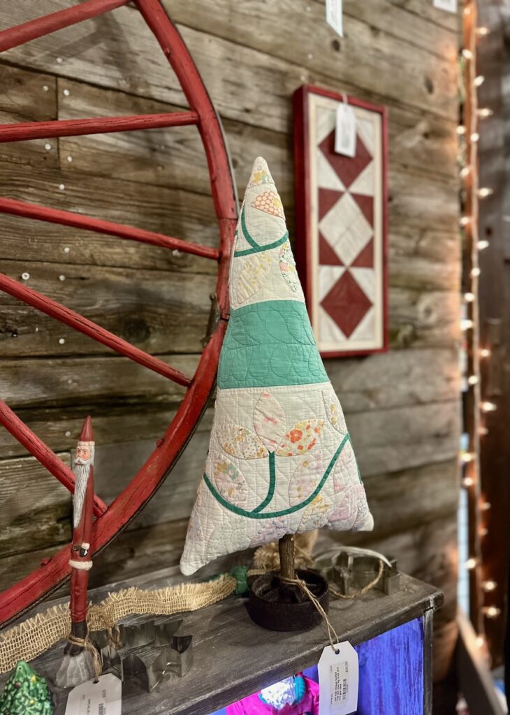 This is a Christmas tree made out of an old quilt, chair leg, and iron gear sitting by skinny Santa, red wagon wheel and fariy lights to show sustainable christmas decorating.