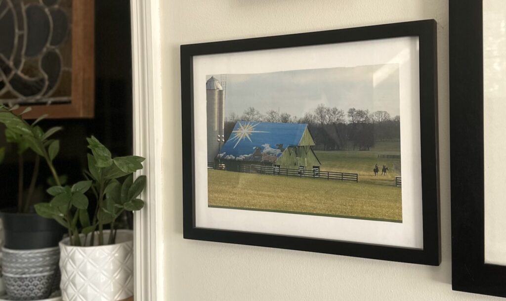 This is a calendar photo of a barn in Brentwood, TN with a beautiful Christmas farm scene on the roof of the barn and two people riding horses in the pasture to show eco friendly christmas star. It's framed on the wall by other frames, plants, and stained glass.