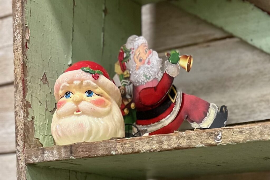 These are two Santa figurines sitting side-by-side on a wooden shelf to show wooden christmas decorations.