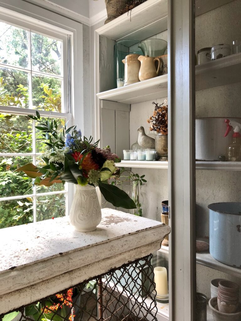 This is a pretty garden shed with a floral bouquet in a pitcher and vases sitting on shelves to show eco friendly things at home.