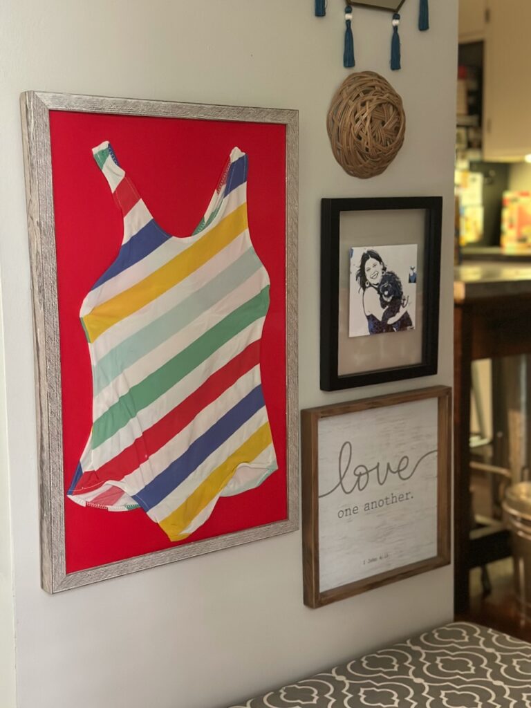 This is a framed swimsuit that's rainbow striped next to a "love one another" Bible verse and a picture of Kat and her pup to show cheap mothers day gifts.